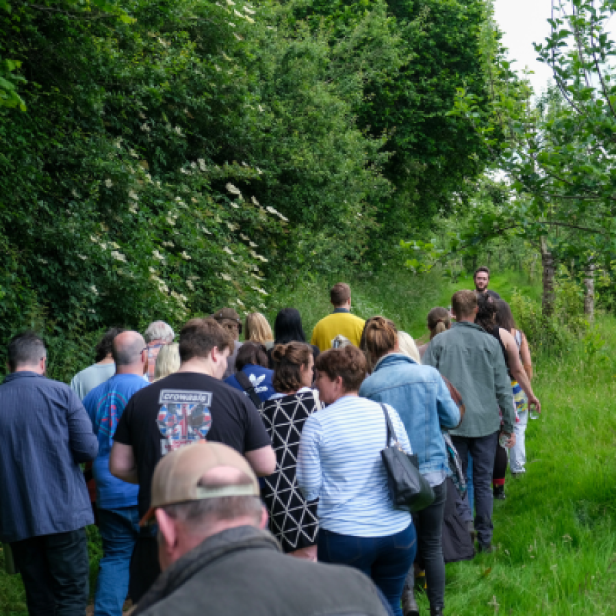 A group of about 19 people taking part in an orchard walk, the leader of the walk is facing the group whose backs are turned to the camera. Long grown grass and a mixture of vibrant green trees fill their surroundings © Rebecca Fletcher Photography